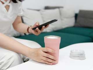Obraz na płótnie Canvas Young woman sitting on sofa looking at her mobile phone with one hand holding a pink cup full of milky drink, focused on her hand and cup.