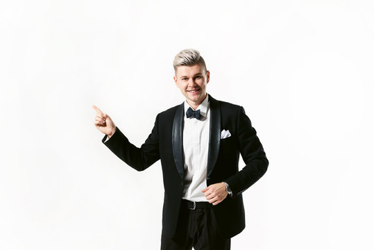 Portrait of young smiling handsome man in tuxedo stylish black suit, studio shot isolated on white background. Showman or toastmaster in jacket with bowtie. He points at copy space by hand