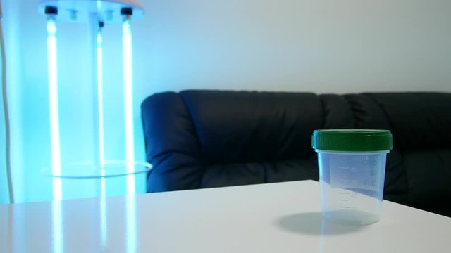 A man puts an empty container for sperm on the table. Analysis of semen. In the corner a bactericidal lamp (ultraviolet) blue color. Artificial insemination concept 4k