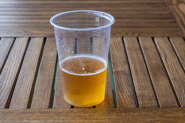 Beer with white foam in a plastic transparent cup on a wooden table.