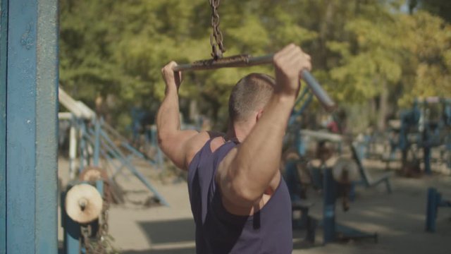 Determined muscular bodybuilder doing lat pulldown heavy weight exercise for back with bar pulled behind neck during outdoor workout. Athletic fit man performing strength exercise for upper back.