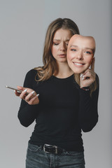 Portrait of a sad girl with a smartphone in one hand and a smiling mask in the other hand.
