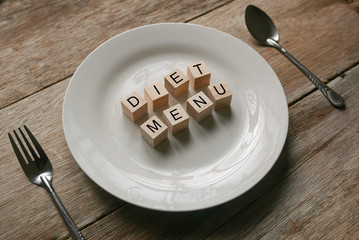 Wooden cube written with 'DIET MENU' on white plate with spoon and fork on wooden background. Food and diet concept.