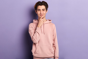 Portrait of a handsome Chinese young man in stylish pink hoodie touching his lips with one finger, thinking expression, isolated on blue background.idea, plan, gay flirting while looking at the camera