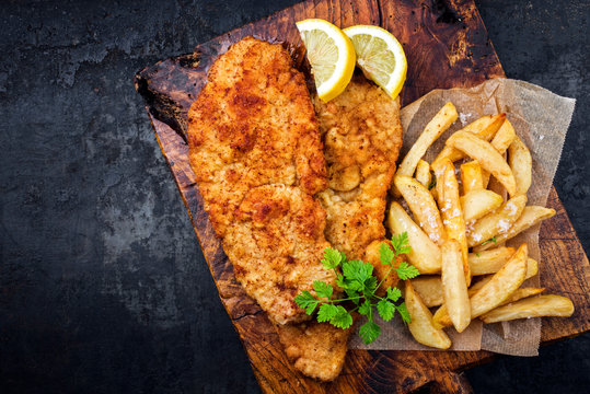 Frittiertes Wiener Schnitzel vom Kalb von der Oberschale mit Pommes Frites und Zitrone als Draufsicht auf einem alten rustikalen Schneidebrett mit Textfreiraum links