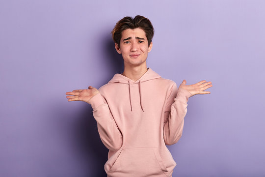 Closeup portrait of puzzled clueless young man wearing pink sweatshirt, arms out asking why what's the problem who cares so what, I don't know. Isolated on blue background