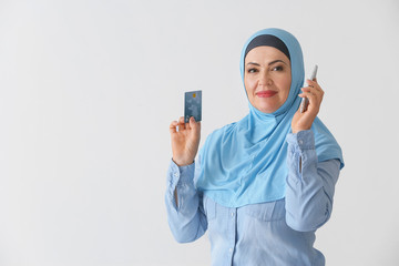Beautiful mature Muslim woman with credit card and mobile phone on light background