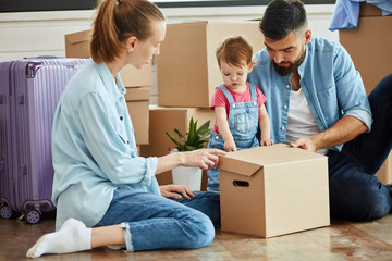 Fototapeta na wymiar Kid wear denim overall, blonde woman wear blue shirt, jeans and white socks and bearded dark-haired man smile and play with box. Background moving boxes and suit