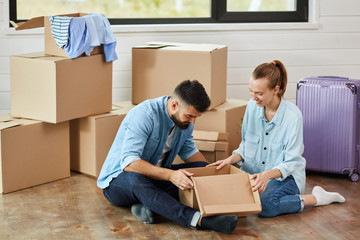 Couple wear blue shirt sit on floor, look into box, smile. Background moving boxes, suit and flower in pot