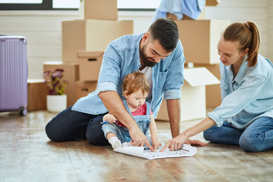 Caucasian family of three sit on floor in new house and look at house plan. Behind them open moving boxes and suitcase.