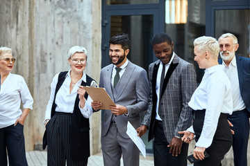 Group of businessmen outdoor. In centre caucasian bearded man in stylish grey suit smile, keep papier. Near african man in checked jacket smile, keep papier. Near them three blonde adult women and man