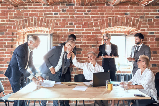 Group of businessmen congratulate colleague with good decision in office. Adult caucasian man wear blue suit kiss hand of aduld blond woman dress white shirt. She smile, look at him