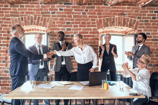 Group of businessmen congratulate colleague with good decision in office. Adult caucasian man wear blue suit handshake aduld blond woman dress white shirt. She smile, look at him. Others clap