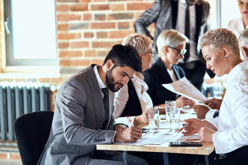 Group of stylishly dressed businessmen sit at table in office and discuss new project. Bearded brunette caucasian man wear gray suit make notes focused