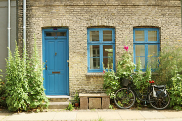 Fototapeta na wymiar An old, gray house with a blue door and blue window frames. Hollyhock and dill grow near the wall.The bike is parked on the pavement. Bench and flower pot on the sidewalk.
