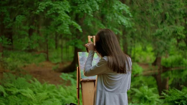 Rear view of a young artist woman painting a landscape in a summer forest. Attractive slender brunette girl draws a picture. Professional in art. Cloudy lighting