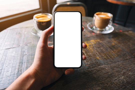 Mockup image of a hand holding and showing black mobile phone with blank white screen and coffee cup on wooden table