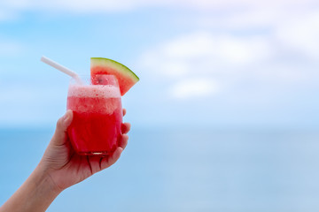 Closeup image of a hand holding a glass of watermelon smoothie by the sea with blue sky background