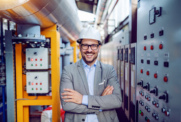 Smiling handsome caucasian businessman in gray suit and with helmet on head standing with arms folded next to dashboard. Power plant interior.
