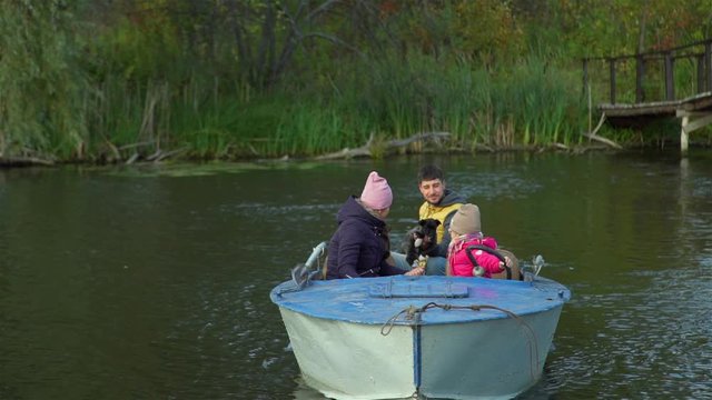 Young Man with his Family Holding Cute Puppy in hus Hand and Driving a Boat on a Lake in Autumn Day. Leisure, Wild Nature and People Concept