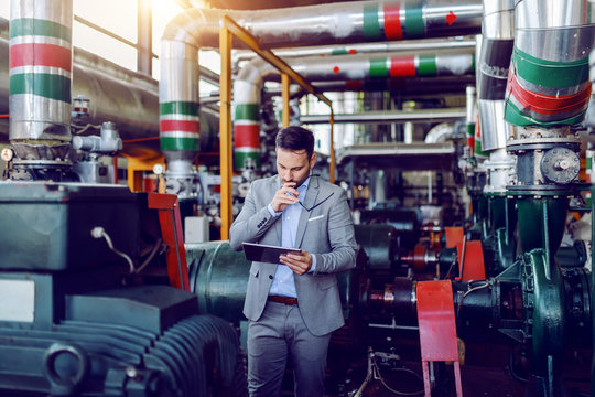 Handsome caucasian manager in gray suit and with eyeglasses in hands using tablet while standing in energy plant.