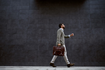 Full length of stylish caucasian designer in gray sweater holding leather bag while passing by gray...