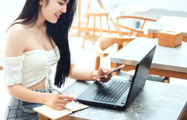 Sexy asian girl smiling and using smartphone with laptop working and writing on notebook in cafe