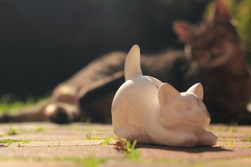 A white cat-shaped penny bank with a grey cat and a garden in the backround.