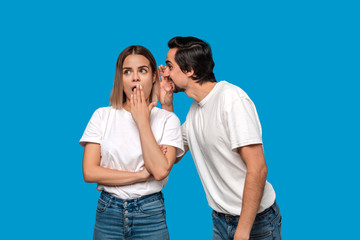 Bearded man with mustaches in white t-shirt tells secret to a young blond girl in white t-shirts and blue jeans standing isolated over blue background. Concept of secret telling.