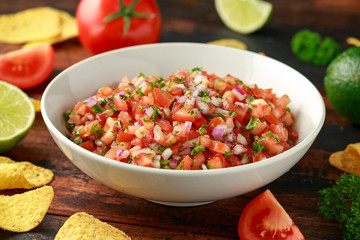 Mexican Tomato Salsa in white bowl with lime, red onion, jalapeno pepper, parsley and tortilla chips on wooden table