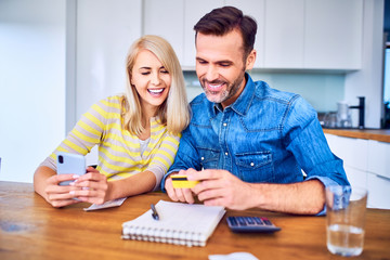Obraz premium Cheerful couple sitting at home together making payments online with smartphone and credit card