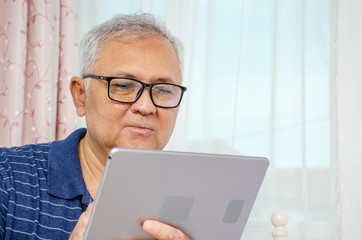 Portrait of senior man sitting at sofa at home and reading news on digital tablet.