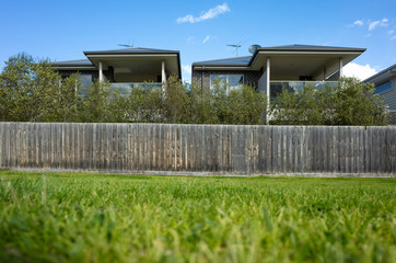 Two-storey residential houses with wooden fence facing vacant land with green grass. VIC Australia. Modern Australian homes in suburb. Copy space for text.