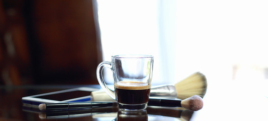 Cup of coffee with the phone and make-up set and brushes on the glass table