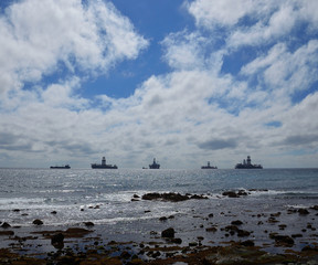 Coast at low tide with stones in foreground, blue sky with clouds and ships anchored in the bay 
