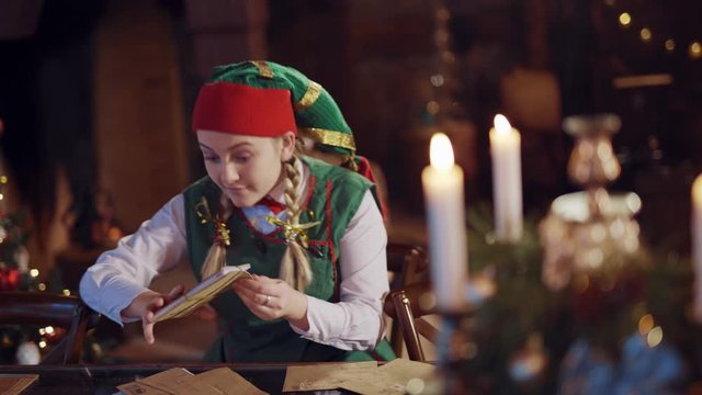 Funny elf with envelopes at the table on festive background. Joyful female elf in green and red costume looking at envelopes for Santa and dances happily.