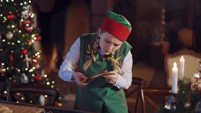 Funny elf holding stack of envelopes for Santa. Dispersed young elf with letters. Envelopes falling on the table from girl's hands on the Christmas tree background.