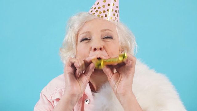 Happy mature childish careless woman in party hat making a noise in the party. Isolated blue background. Celebration, happiness. Lifestyle