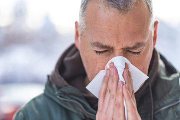 Closeup portrait of mature man with allergy or cold, blowing his nose with a tissue, looking...