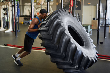 Naklejka na ściany i meble Full length side view of muscular African man flipping tire during cross workout in modern gym, copy space