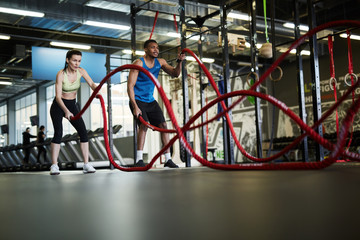 Wide angle portrait of fit young couple exercising with battle ropes during strength workout in cross functional gym, copy space