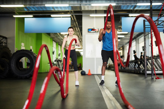 Full length portrait of fit couple exercising with battle ropes during strength workout in modern gym, copy space