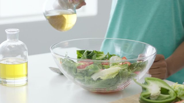 Woman preparing fresh salad in glass salad bowl, pouring oil over lettuce leaves and mixing with salad servers