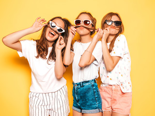 Three young beautiful smiling hipster girls in trendy summer clothes. Sexy carefree women posing near yellow wall in studio. Positive models going crazy and having fun.Making mustache with hair