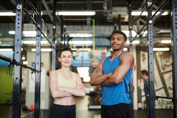 Waist up portrait of sportive couple, African man and Caucasian woman, posing confidently standing in modern gym and smiling at camera
