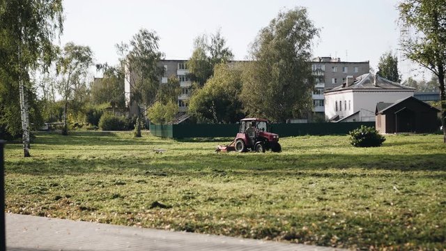 Tractor rides through the Park and mows the lawn with a lawn mower. View from afar. Territory cleaning.