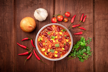Chili con carne, a Mexican stew with red beans, meat, and chili peppers, shot from the top on a dark rustic wooden background with ingredients