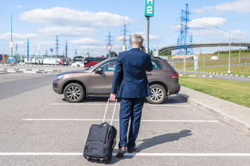 Man in a blue suit with a suitcase walking to his car and calling by phone at the airport parking. Business trip concept