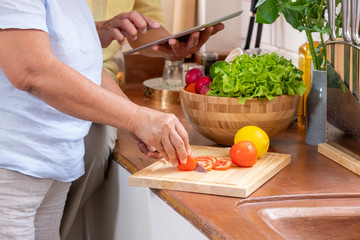 Asian senior wife slicing tomatoes on wooden cutting  board and using tablet computer to searching menu recipe.senior with technology lfiestyle.aging at home