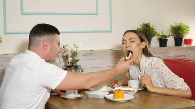 young man feeds smiling girlfriend with tasty berry cake having date at wooden table in confectionery shop